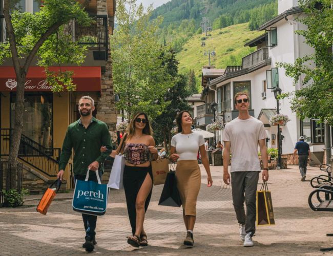A sunny alpine street with friends walking and carrying shopping bags, framed by trees and cable cars in the mountains.