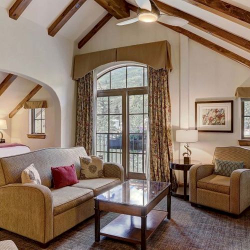 A cozy mountain-style living room with vaulted wooden beams, large windows, a sofa and chairs, and a wall-mounted TV.