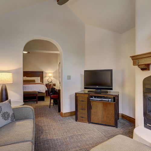 A hotel living room with a TV on a wooden cabinet, a sofa, a fireplace, and an archway leading to a bedroom.