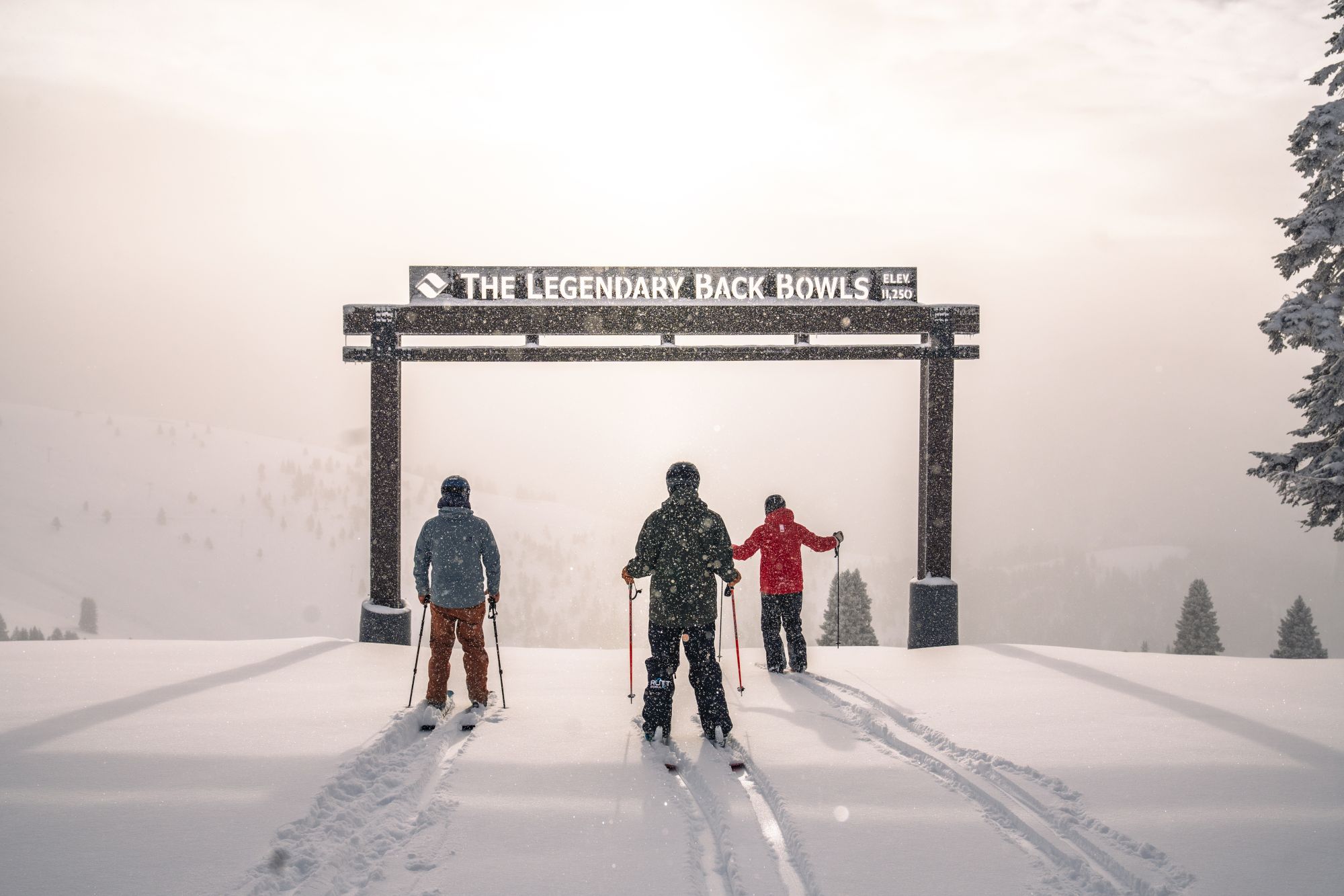 Skiers trekking in snowy mountains under an arch reading &ldquo;THE LEGENDARY BACK BOWLS,&rdquo; with falling snow all around.