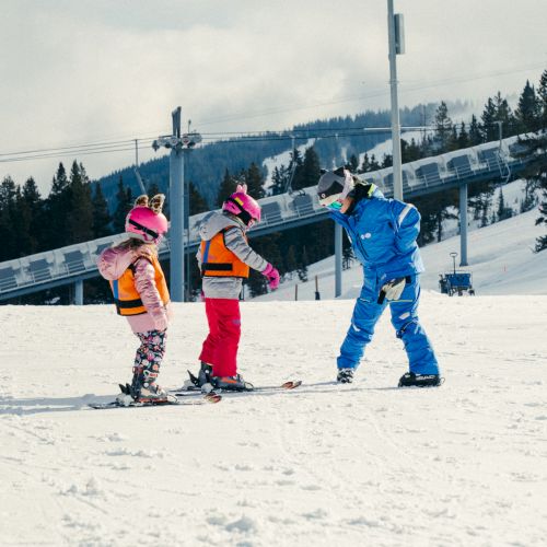 Three kids in winter helmets and colorful ski gear stand on a snowy slope near ski lifts, taking a fun moment outdoors.