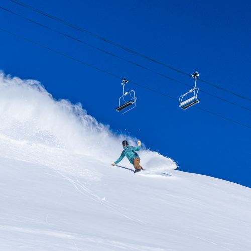 A skier carving through fresh snow on a steep slope while overhead ski-lift chairs glide against a bright blue sky.