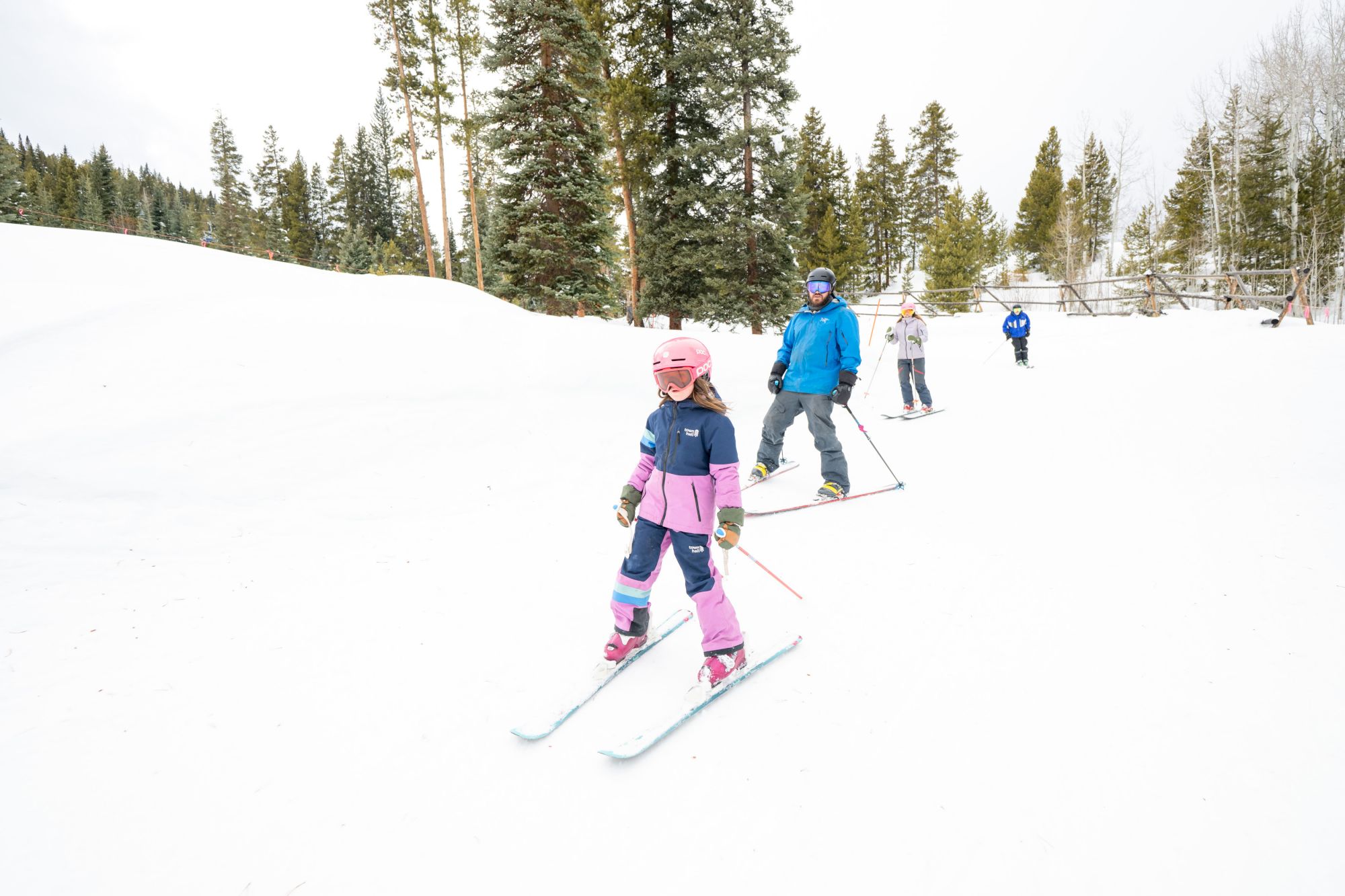 Skiers glide on a snowy run in colorful jackets and helmets under pine trees.
