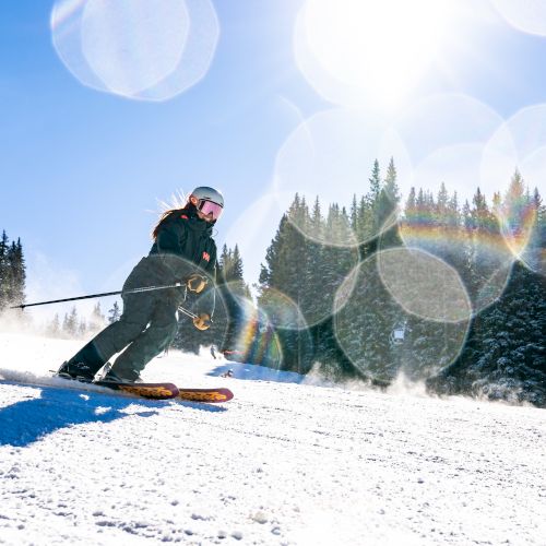 A skier carves through fresh snow in a pine forest, with bright sun flares and lens-bubble rings in the sky.