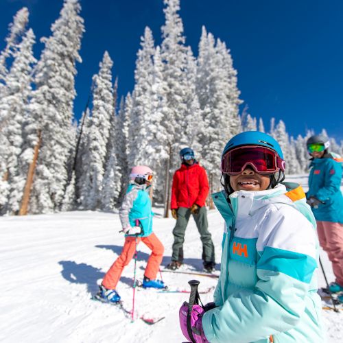 Kids ski on a sunny snowy slope, wearing helmets and goggles, with tall pine trees in the background.
