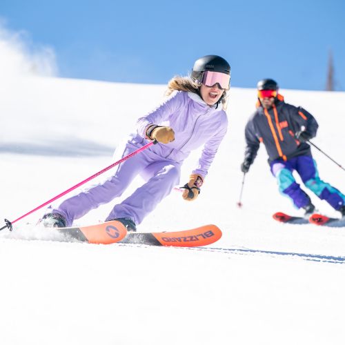 A skier in a purple suit carves down a snowy slope, kicking up powder while another skier follows behind.