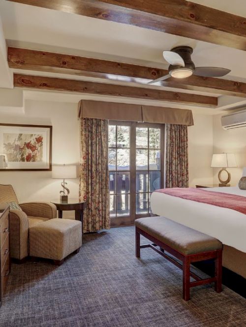 A bright hotel bedroom with a wooden ceiling fan, bed, curtains, window, air conditioner, and a small dresser with TV.