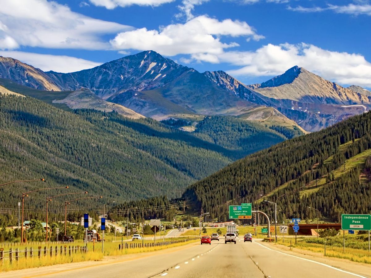 Cars drive on a mountain highway in a forested valley under bright clouds.