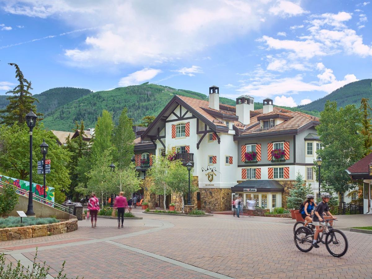An Alpine village walkway with a white chalet hotel, people strolling, cyclists riding by, and green mountains under sunny clouds.