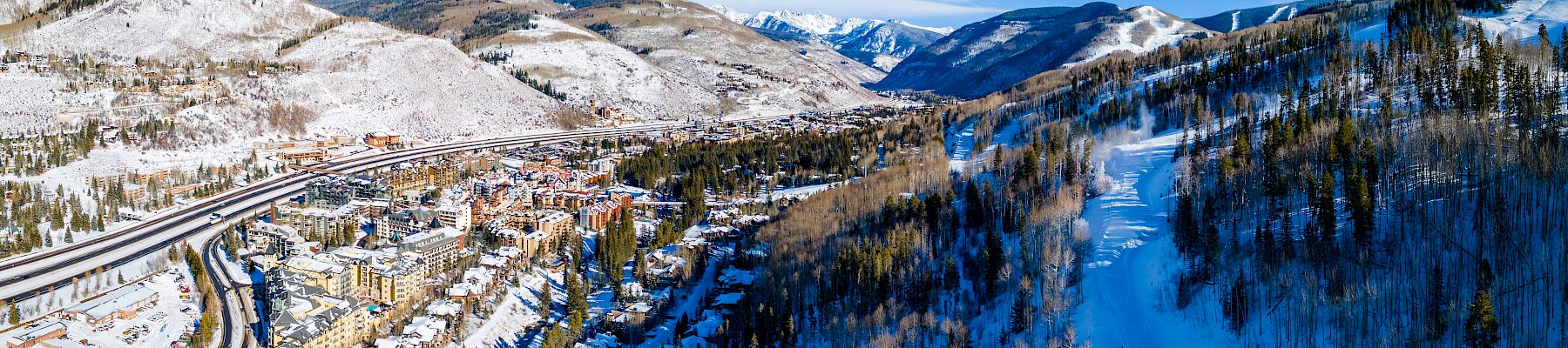 Snowy mountain valley with a ski resort village, winding roads, pine forest, and a frozen-blue river under a clear sky.