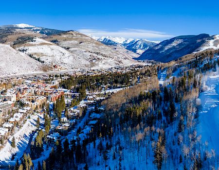 Snowy mountain valley with a ski resort village, winding roads, pine forest, and a frozen-blue river under a clear sky.