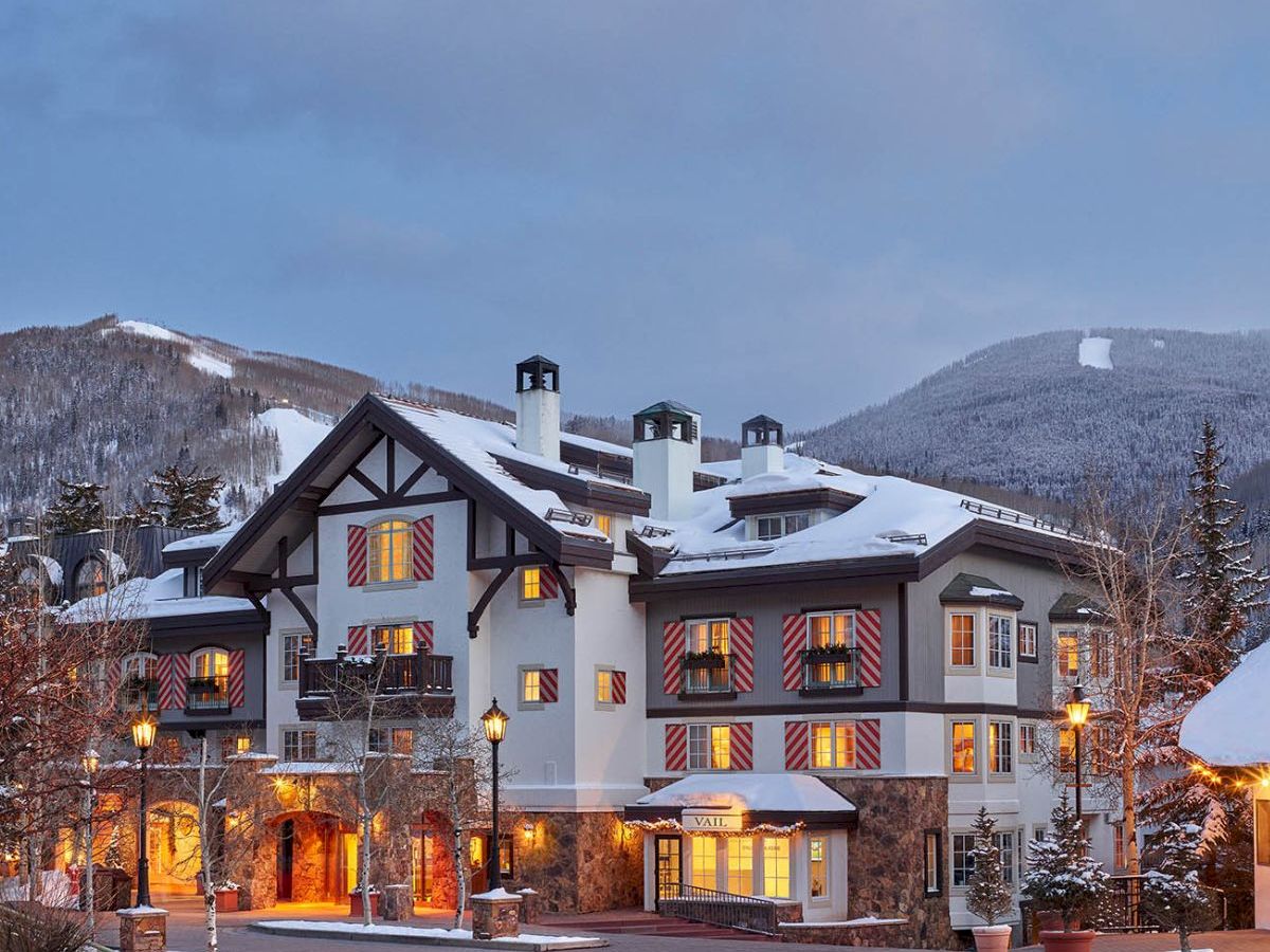 A snow-covered alpine lodge with lit windows sits in a valley under a blue sky between winter mountains.