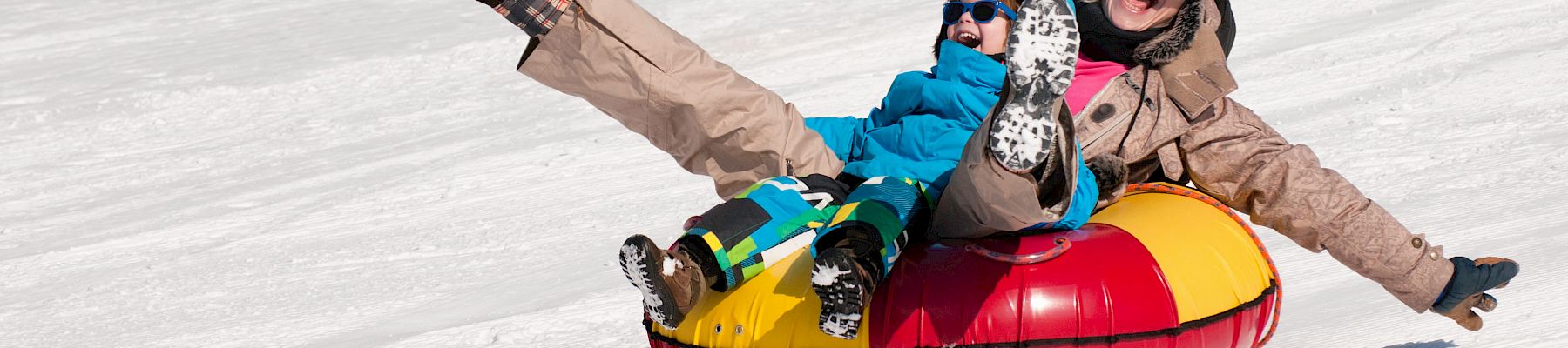 Two people in winter gear tumble down snowy slopes while snowtubing in a bright inflatable tube, laughing and cheering.