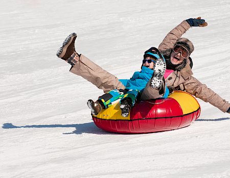Two people in winter gear tumble down snowy slopes while snowtubing in a bright inflatable tube, laughing and cheering.