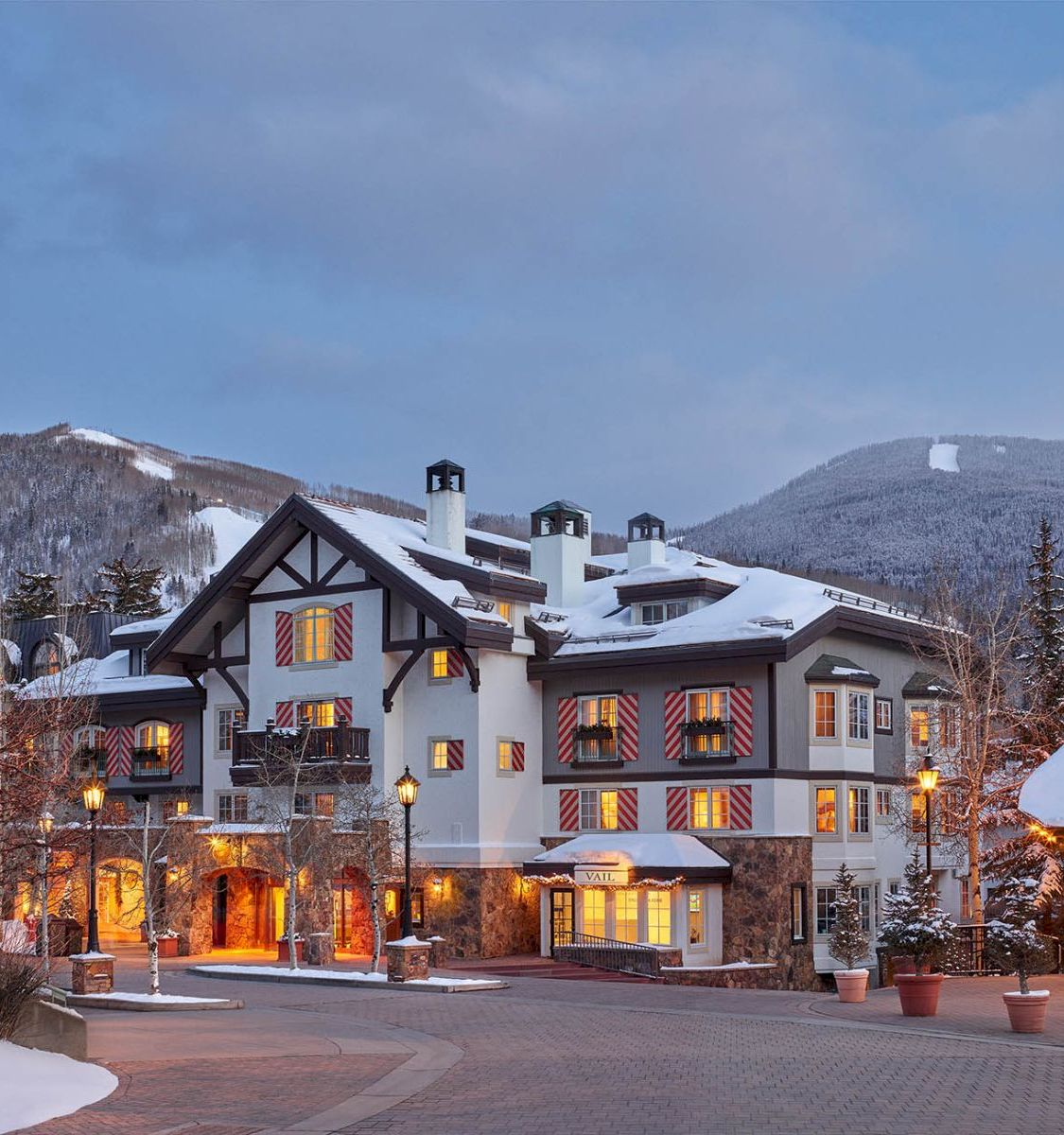 A snowy alpine chalet resort with warm windows and red-white shutters, set against wintry mountains under a blue sky.