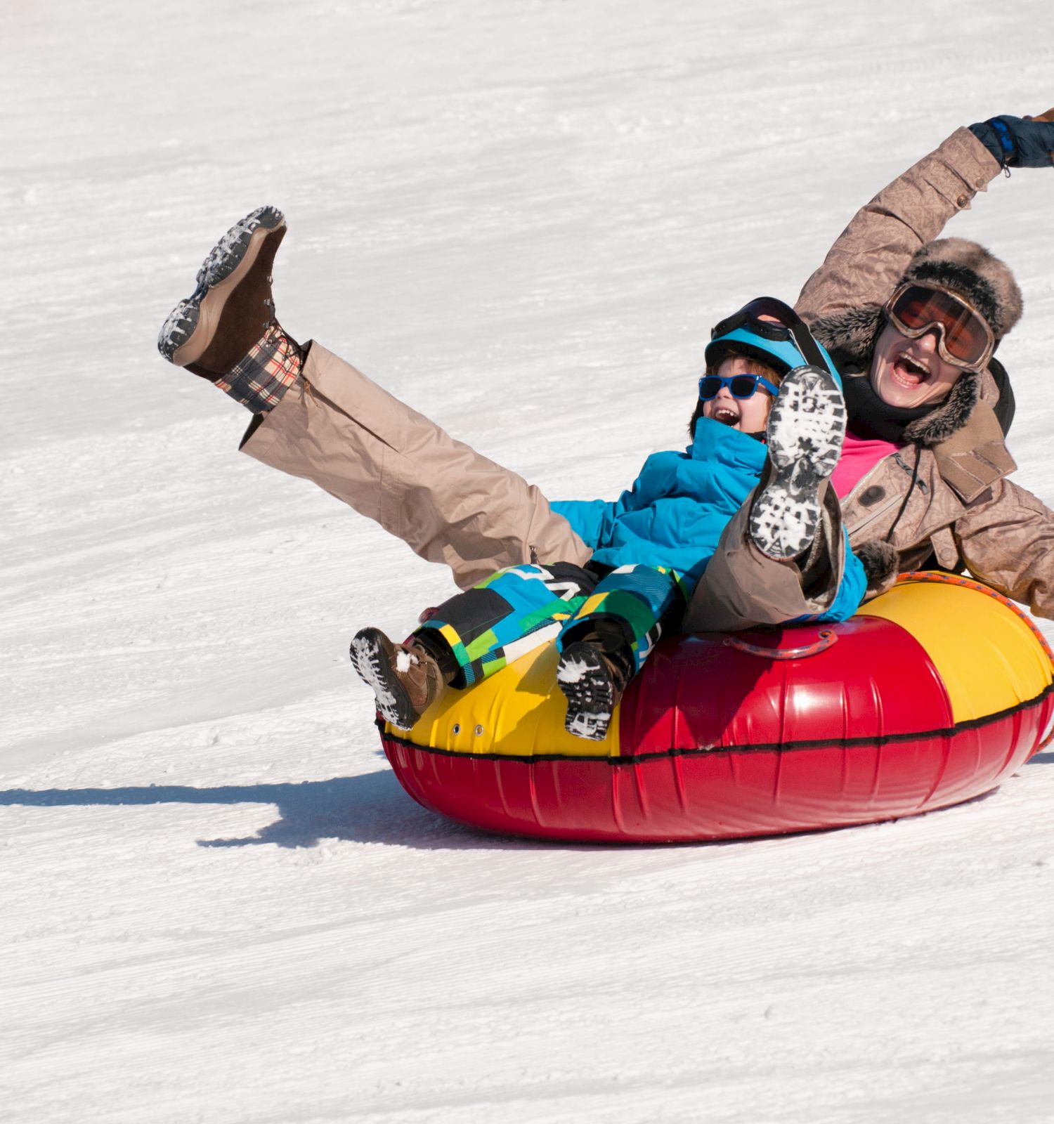 Two people laugh while sliding down a snowy hill in a red/yellow inner tube, wearing winter gear and goggles.